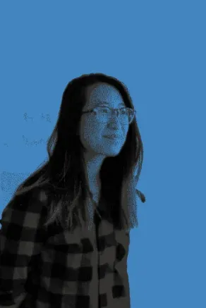 A smiling woman with long dark hair and thick glasses wearing a flannel shirt.